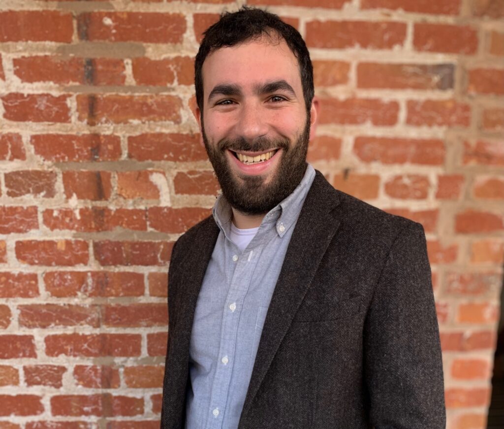A man with short dark hair and a beard smiles while standing in front of a red brick wall, wearing a gray blazer over a light blue shirt.