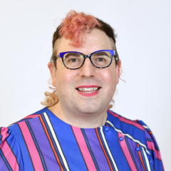 A person with pink curly hair, glasses, and a bright smile wears a blue and purple striped top, posing against a plain white background.