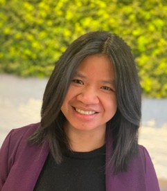 A woman with straight dark hair, wearing a purple blazer and black top, smiles in front of a blurred green and yellow leafy background.