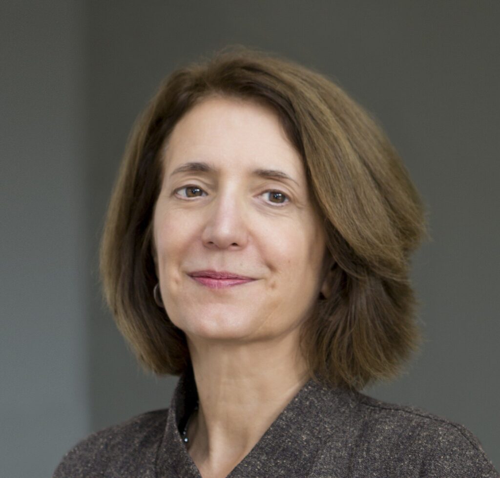 A woman with shoulder-length brown hair wearing a dark textured jacket and black top, standing against a plain gray background, looking at the camera with a slight smile.