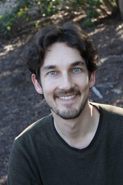 A man with wavy brown hair and a short beard smiles at the camera outdoors, wearing a dark green sweater. The background is blurred with soil and green foliage.