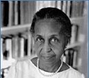 Black-and-white photo of an older woman with short hair, wearing a pleated white blouse and a pearl necklace, standing in front of a blurred background of bookshelves.
