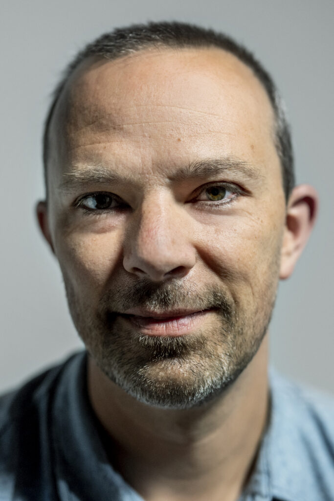 A middle-aged man with short hair, light beard, and mustache looks directly at the camera, wearing a light blue collared shirt against a plain background.