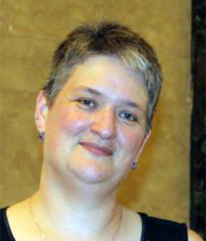 A person with short, grayish hair and light skin smiles gently, wearing a dark top and earrings, standing in front of a tan and dark marbled wall.