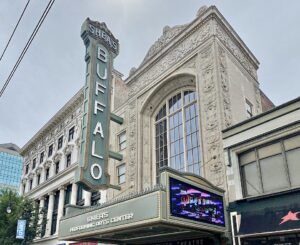 A historic theater with ornate architecture, a tall vertical sign reading Sheas Buffalo, and a digital marquee above the entrance displaying event information.