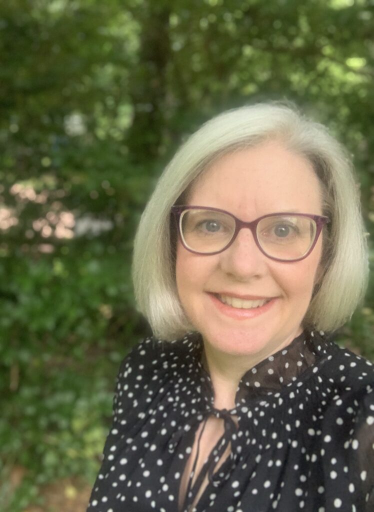 A woman with gray hair and glasses smiles outdoors, wearing a black blouse with white polka dots. Green foliage is blurred in the background.